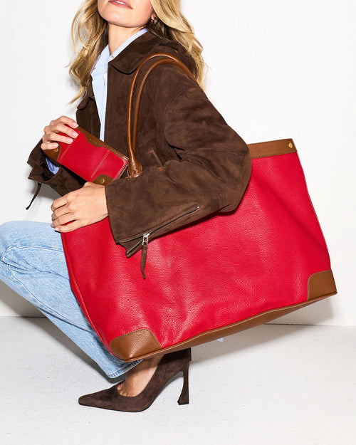 A woman in a brown suede jacket, light blue shirt, and jeans carries a large red leather tote bag with brown leather trim and a matching red pouch.