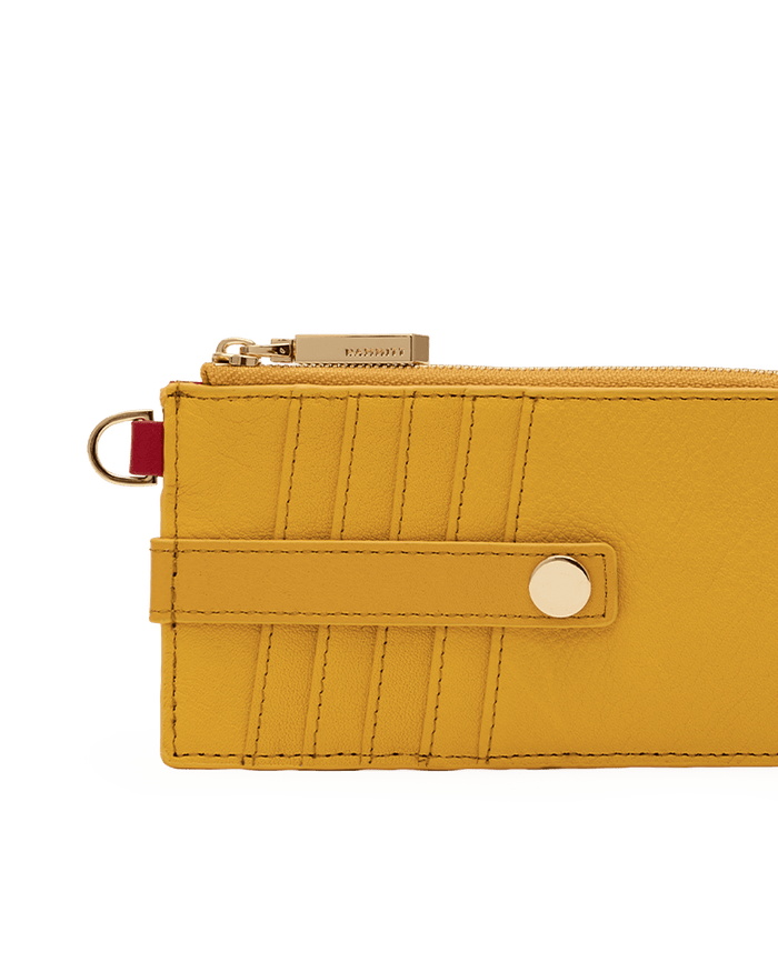 A close-up of a mustard yellow leather zip-around wallet or card holder with gold-tone hardware, a snap strap closure, and a small red accent ring on a plain white background.