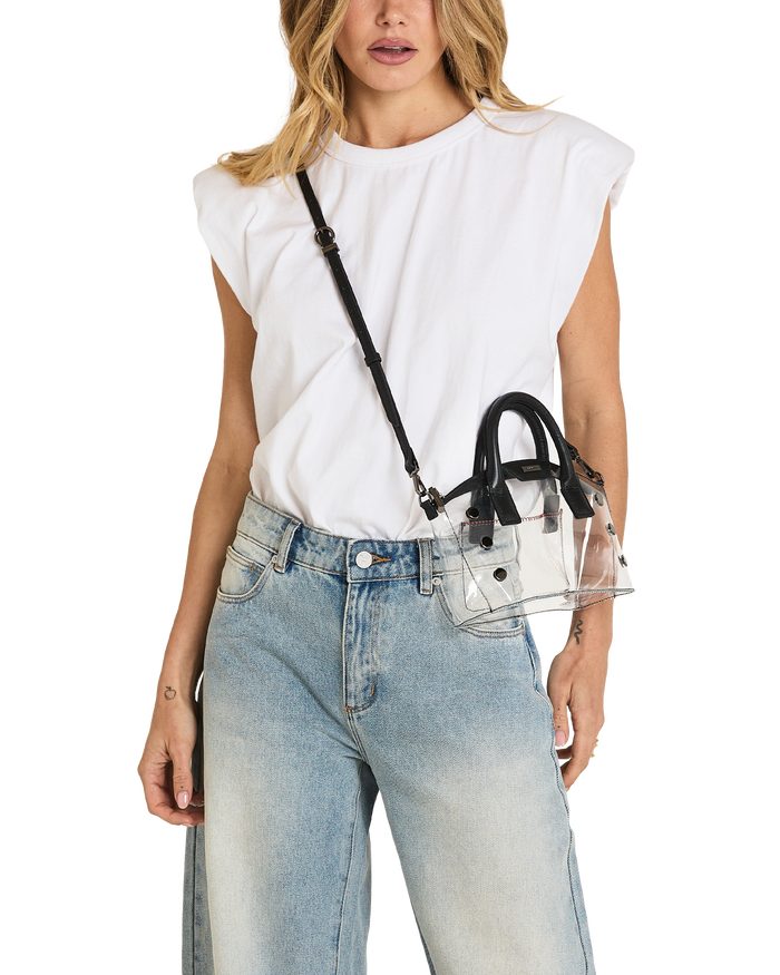 A woman wearing a white sleeveless top and light blue jeans carries a small clear handbag with black handles and a black shoulder strap against a plain white background.