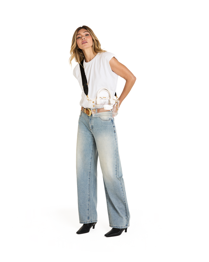 A woman modeling a small clear handbag with white details and gold hardware wearing a white sleeveless top, light-wash wide-leg jeans, a gold belt, and black ankle boots against a plain white background.