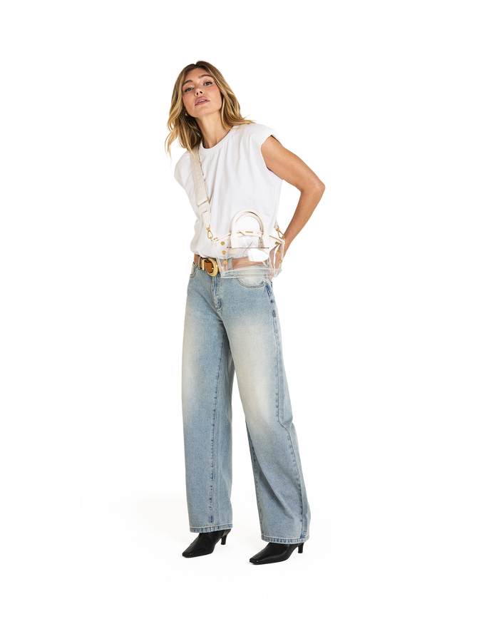 A woman modeling a white sleeveless top, light-wash wide-leg jeans, a gold belt, and black pointed-toe heels, while carrying a small white handbag with a gold chain strap.